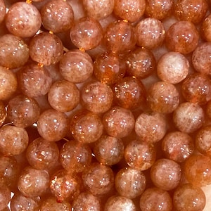 Può includere: Una collezione di perline rotonde in pietra del sole caratterizzate da una calda tavolozza di colori arancione e pesca con inclusioni interne naturali che creano un effetto scintillante e riflettente sulle superfici lisce e levigate.