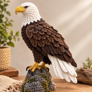 Puede incluir: Una figura de águila calva tejida a ganchillo con cabeza y cola blancas, alas y cuerpo marrón oscuro, y pico y garras de color amarillo brillante. El águila está posada sobre una base de roca tejida en gris con detalles verdes.