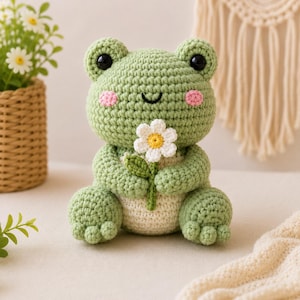 May include: A light green crocheted frog plush toy with a cream-colored belly, black eyes, and pink cheeks. The frog is holding a small white daisy flower with a yellow center and a green stem in its hands, featuring a textured yarn construction.