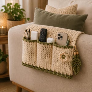 May include: A cream-colored crochet sofa armrest organizer featuring three deep pockets with dark green trim. The organizer is adorned with a small crocheted flower and a dangling leaf-shaped charm with wooden beads, holding various household items.