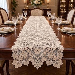Puede incluir: Un camino de mesa de encaje beige, largo y rectangular, con un intrincado patrón de flores y hojas y bordes festoneados, colocado sobre una mesa de comedor de madera oscura con platos blancos, servilletas y cristalería.