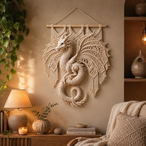 Könnte beinhalten: Ein cremefarbener Makramee-Wandbehang mit einem detaillierten Drachendesign, kunstvoll geknoteten Flügeln und einem geringelten Schwanz, der an einem horizontalen Holzstab befestigt ist und komplexe Handwerkskunst zeigt.