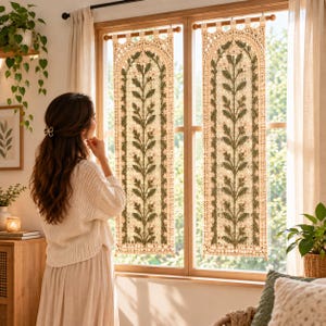 Pode incluir: Duas cortinas de janela retangulares em macramé com um design de arco no topo. O cordão de cor creme é tecido num padrão de treliça com um motivo de videira vertical central em verde escuro e pequenos detalhes florais rosa claro.