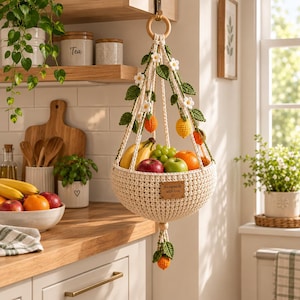 May include: A cream-colored crocheted hanging fruit basket featuring decorative green leaves, white flowers, and small orange and yellow fruit accents. The basket has a wooden ring at the top, a wooden bead at the bottom, and a leather tag labeled handmade with love.