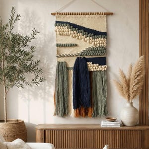 May include: A rectangular woven wall hanging suspended from a wooden rod. The textile features a cream-colored base with horizontal bands of navy blue, sage green, and cream-colored yarn knots, finished with long, hanging fringe in navy, sage, and rust.