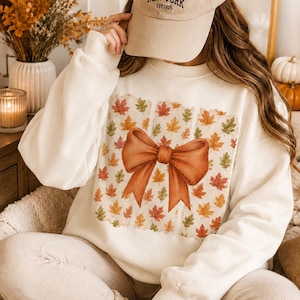 Pode incluir: Uma camisola de gola redonda cor de creme com um grande gráfico central de um laço cor de laranja queimado sobre um padrão de pequenas folhas de outono multicoloridas em tons de vermelho, laranja e amarelo, com mangas compridas.