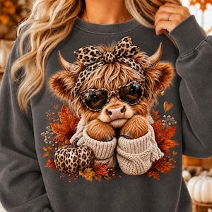 May include: A dark gray crewneck sweatshirt featuring a graphic of a Highland cow wearing heart-shaped sunglasses and a leopard print bow. The cow is depicted in a beige knit sweater surrounded by orange autumn leaves and a leopard print pumpkin.