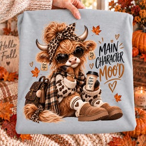 May include: A light grey sweatshirt featuring a graphic of a fluffy highland cow wearing a leopard print headband, sunglasses, a plaid scarf, and tan boots. The cow holds a coffee cup with the text More Coffee Please and Main Character MOO'D.