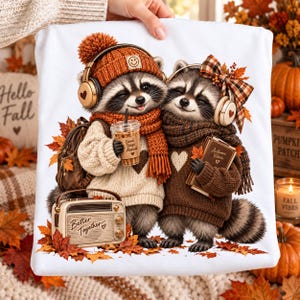 May include: A white t-shirt featuring a graphic of two raccoons wearing sweaters, scarves, and headphones. One raccoon holds an iced coffee cup labeled Good Days Ahead, while the other holds a book labeled Autumn Vibes. A vintage radio labeled Better Together sits at their feet amidst fallen autumn leaves.