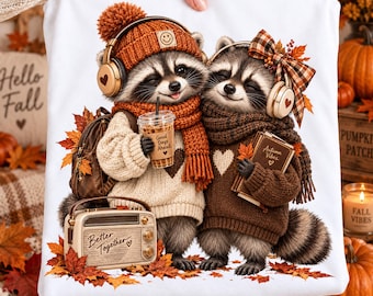 May include: A white t-shirt featuring a graphic of two raccoons wearing knit sweaters, scarves, and headphones. One raccoon holds a coffee cup labeled Good Days Ahead, while the other holds a book labeled Autumn Vibes. A vintage radio labeled Better Together sits at their feet, surrounded by orange autumn leaves.