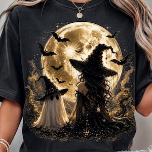 May include: A charcoal gray t-shirt featuring a graphic print of a witch in a black dress and pointed hat standing next to a white ghost. The scene is set against a large, glowing full moon with several black bats flying in the night sky.