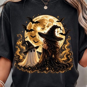 May include: A black t-shirt featuring a graphic print of a witch in a black dress and pointed hat sitting next to a white ghost in a pointed hat. The scene is set against a large, glowing yellow full moon with flying bats and golden sparkles.