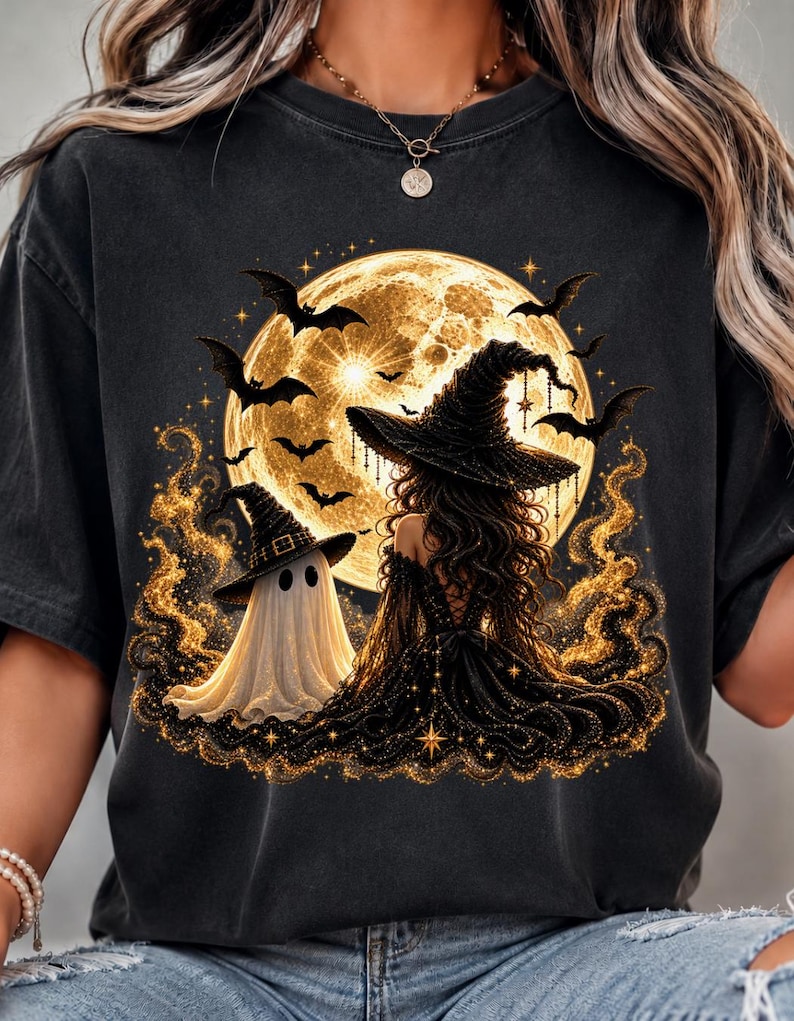 May include: A black t-shirt featuring a graphic print of a witch in a black dress and pointed hat sitting next to a white ghost in a pointed hat. The scene is set against a large, glowing yellow full moon with flying bats and golden sparkles.
