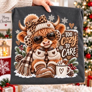 May include: A dark grey t-shirt featuring a graphic of a fluffy highland cow wearing a leopard print bow, earmuffs, and sunglasses. The cow wears a scarf and boots, holding a mug of cocoa. Text reads: Too Cozy To Care, with snowflakes.