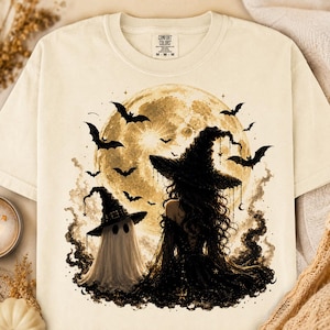 May include: A cream-colored t-shirt featuring a graphic of a witch and a ghost standing before a large, full moon. The design includes several flying bats, dark swirling smoke, and small star accents. The shirt has a Comfort Colors brand label.