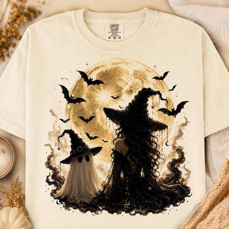 May include: A cream-colored t-shirt featuring a graphic of a witch and a ghost standing before a large, full moon. The design includes several flying bats, dark swirling smoke, and small star accents. The shirt has a Comfort Colors brand label.