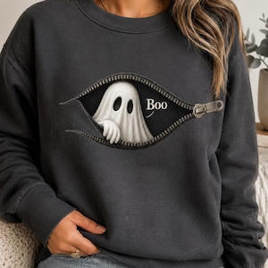 Puede incluir: Una sudadera de cuello redondo gris carbón con un gráfico de un fantasma blanco asomándose detrás de una cremallera metálica parcialmente abierta. El fantasma tiene dos ojos ovalados oscuros y una mano visible, con la palabra Boo en blanco.