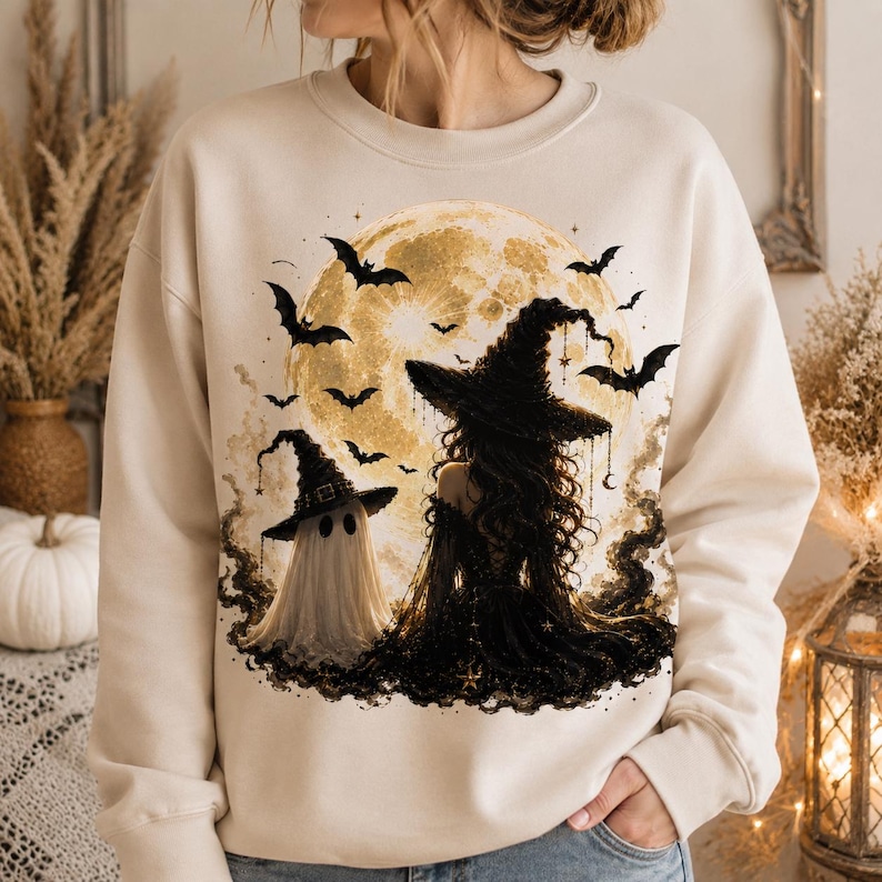 May include: A cream-colored crewneck sweatshirt featuring a graphic print of a ghost wearing a witch hat standing next to a witch with long dark hair and a pointed hat, all set against a large yellow full moon with flying black bats.