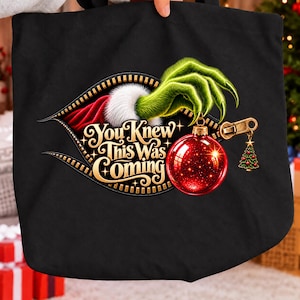 May include: A black tote bag featuring a graphic of a green furry hand emerging from a zip opening. The hand holds a shiny red Christmas bauble. The text reads You Knew This Was Coming in gold lettering, accompanied by a small Christmas tree charm.