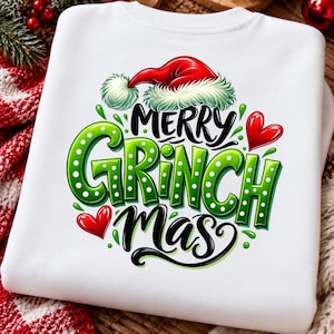 May include: A white sweatshirt featuring a graphic print with the text Merry Grinch Mas in black and green lettering. The design includes a red Santa hat with white fur trim, two red hearts, and green polka-dot accents on the letters.