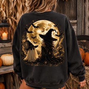 May include: A charcoal gray long-sleeve sweatshirt featuring a large back graphic of a full moon, a witch in a pointed hat, and a ghost wearing a small hat. The scene includes several flying bats and golden sparkles against a dark, textured background.