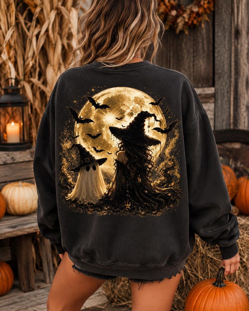 May include: A charcoal gray long-sleeve sweatshirt featuring a large back graphic of a full moon, a witch in a pointed hat, and a ghost wearing a small hat. The scene includes several flying bats and golden sparkles against a dark, textured background.