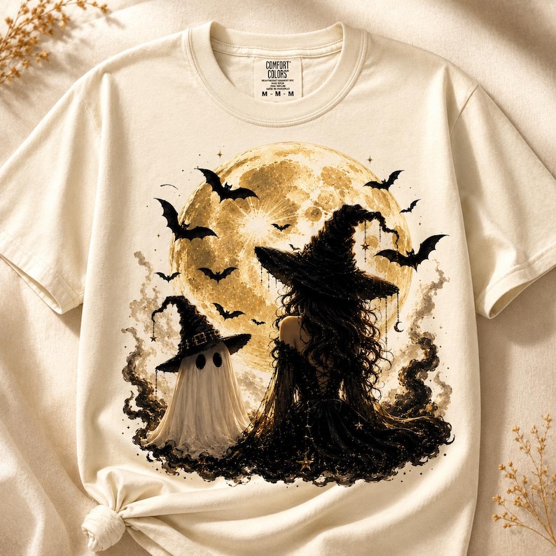 May include: A cream-colored short-sleeve t-shirt featuring a graphic print of a person in a black witch hat and dress standing next to a ghost in a witch hat. The background shows a large yellow moon, black bats, and dark swirling smoke.