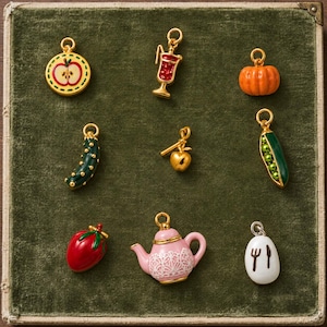 May include: A collection of nine gold-toned metal charms featuring enamel details, including an apple slice, a red drink with a straw, an orange pumpkin, a green cucumber, a gold apple, a green pea pod, a red strawberry, a pink teapot, and a white egg.