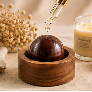 May include: A dark brown, spherical object sits inside a circular wooden base with a visible grain pattern. A clear glass dropper with a gold-colored top is positioned above the sphere, releasing a single droplet of liquid onto its smooth surface.