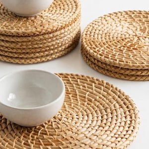 Peut inclure: Une collection de sets de table ronds en rotin tressé de couleur marron clair naturel. Les sets de table présentent un motif circulaire en spirale. Deux petits bols en céramique blanc cassé lisses sont posés sur deux piles de ces sets.