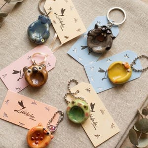 Può includere: Sei portachiavi in ceramica a forma di animale nei colori blu, marrone, giallo, verde e arancione, ciascuno attaccato a un'etichetta del marchio Libélula. I portachiavi presentano piccole figure arrotondate con dettagli del viso distinti.