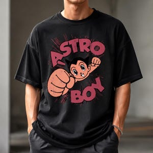 May include: A black short-sleeve t-shirt featuring a graphic print of the character Astro Boy with a raised fist. The design includes the text "ASTRO BOY" in large, distressed red lettering arched above and below the character illustration.