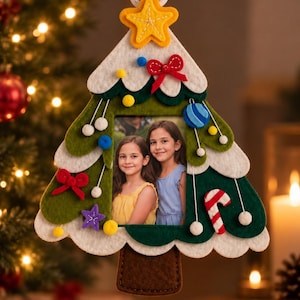 Puede incluir: Un marco de fotos de fieltro con forma de árbol de Navidad que presenta capas de tela verde y blanca, decorado con pequeños lazos, estrellas y un bastón de caramelo de fieltro. Una abertura central rectangular muestra una foto de dos niños.
