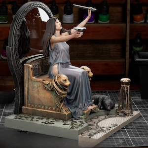 Pode incluir: Uma estatueta de resina pintada de uma mulher com cabelos longos e escuros vestindo um vestido cinza fluido, sentada em um trono cor de bronze com apoios de braço em forma de cabeça de leão. Ela segura uma tigela e uma varinha prateadas.
