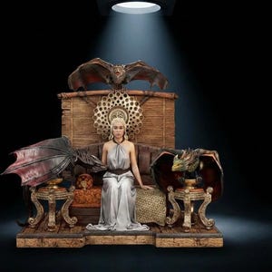 May include: A detailed figurine set featuring a central figure with blonde hair in a silver dress seated on a wooden throne. Two winged dragon figures are positioned on ornate stands on either side, with a third dragon perched atop the throne back.