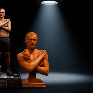 May include: A full-body figurine of a man in black shorts with a white bird on his shoulder standing on a base, alongside a terracotta-coloured bust of the same man holding a small bird against his chest, both illuminated by a single overhead light.
