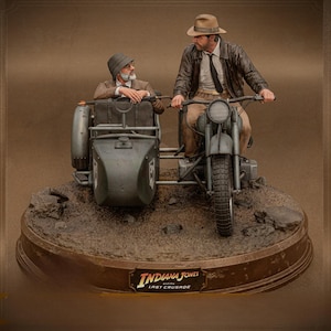 Può includere: Un set di statuette da collezione dettagliate che raffigura due personaggi su una motocicletta d'epoca con sidecar. I personaggi indossano cappelli e abiti d'epoca. La base è strutturata come terra e include una targa con la scritta Indiana Jones and the Last Crusade.