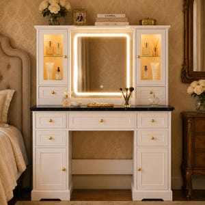 May include: A white vanity table with a black marble countertop, featuring a central LED-lit mirror, two glass-fronted upper cabinets with interior lighting, multiple storage drawers with gold knobs, and lower cabinets on each side for organisation.