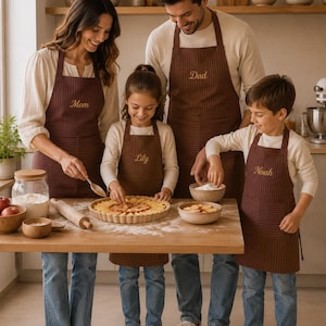Puede incluir: Un conjunto de cuatro delantales marrones a rayas a juego con nombres bordados. Cada delantal incluye un bolsillo frontal y correas ajustables para el cuello. Los delantales están personalizados con el texto Mom, Lily, Dad y Noah en letras doradas.