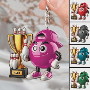 May include: A keychain featuring a pink bowling ball character wearing a backwards baseball cap and sneakers, standing next to a gold trophy with three white bowling pins. The trophy base has the name ALEX printed in black on a yellow background.
