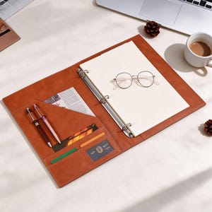 Può includere: Una cartella portadocumenti in similpelle color cuoio con meccanismo a tre anelli in metallo, un blocco note e tasche interne. L'organizer include due penne, uno scomparto trasparente per documenti e una tasca. Un paio di occhiali tondi è appoggiato.