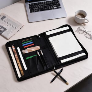 May include: A black leather-like zippered portfolio organizer featuring an interior grid-lined notepad, multiple card slots, document pockets, and two pen loops. The open case displays organized storage compartments for business essentials and stationery.