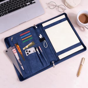 May include: A navy blue zipped portfolio organiser featuring multiple interior slots for cards, a smartphone, pens, and a charging cable. The right side contains a grid-lined notepad secured by a strap, while the left side holds a tablet.