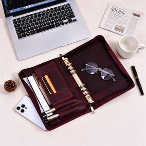 May include: A dark burgundy leather zippered binder featuring a gold-toned six-ring metal mechanism, multiple interior card slots, and two pen loops. The open organizer contains a pair of round-frame eyeglasses resting on the right side panel.