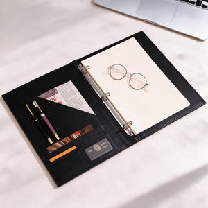 Peut inclure: Un classeur à trois anneaux en similicuir noir ouvert révélant un bloc de papier blanc, une paire de lunettes rondes à monture métallique, deux stylos dans une pochette, une petite carte orange et un porte-carte en plastique transparent.