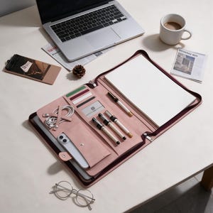 May include: A dusty rose leather portfolio organiser featuring a zipped closure, a large notepad, multiple slots for pens, cards, and a smartphone, along with a dedicated compartment for a tablet or laptop, all arranged in a structured interior layout.