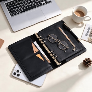 May include: A black leather-like planner organizer with a gold-toned six-ring binder mechanism, featuring internal pockets, a zippered compartment, a pen loop with a black pen, and a pair of round gold-rimmed glasses resting on the open cover.