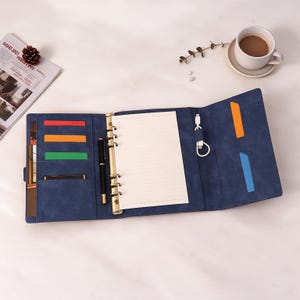 Op de afbeelding: Een blauwe lederlook ringband-organizer met een goudkleurig zes-ringsmechanisme, gelinieerd papier, een zwarte pen en meerdere binnenvakken met rode, oranje, groene en blauwe accenten. Een witte USB-oplaadkabel is aan de rechterkant bevestigd.