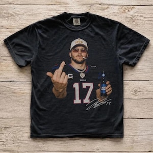 May include: A black short-sleeve t-shirt featuring a graphic print of a person wearing a white MVP cap, sunglasses, and a Buffalo Bills jersey with the number 17. The person is holding a Bud Light bottle and giving the middle finger, with a signature below.