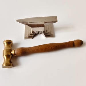 Könnte beinhalten: Ein polierter, silberfarbener Metallamboss mit abgestuftem Sockel hinter einem kleinen Hammer mit zylindrischem Messingkopf und einem glatten, hellbraunen Holzgriff mit abgerundetem Ende, beide auf einer schlichten weißen Oberfläche.